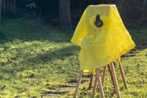 Eine Kiste steht auf 2 Böcken im Garten. Sie ist mit einem gelben Regenponcho abgedeckt. vorne sieht man eine Linse herausragen.