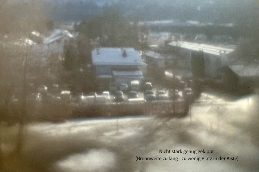 Unscharfes Bild von Häusern, Parkplatz, verschneite Wiese. Vorne und hinten deutlich unscharf, in der Mitte mäßig scharf